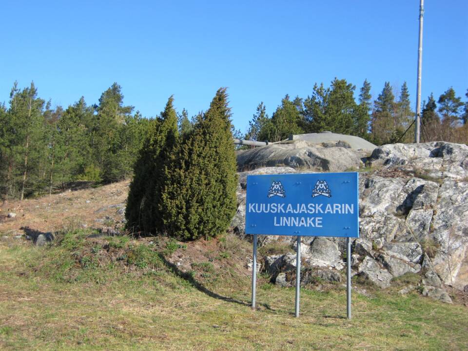 Etualalla ruohikossa suuri kyltti "Kuuskajaskarin linnake", taustalla kalliolla näkyy tykki ennen havumetsää.