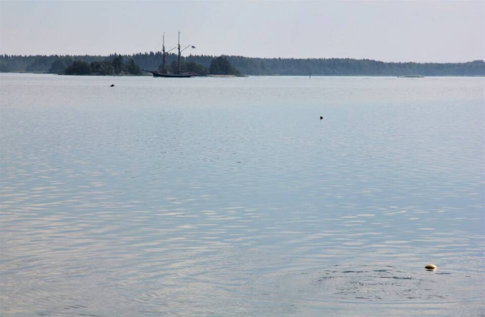 Merimaisema, horisontissa metsäisiä saaria/mannerta, niiden edessä kaksimastoinen purjelaiva.