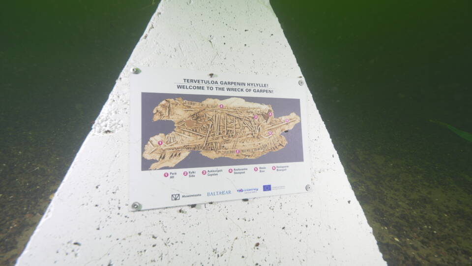 Kolmionmallinen teline, johon kiinnitettynä valokuva/pohjapiirrustus hylystä sen nykyisessä, hajonneessa tilassa, opaskyltti meren pohjassa hylylle sukeltaville.