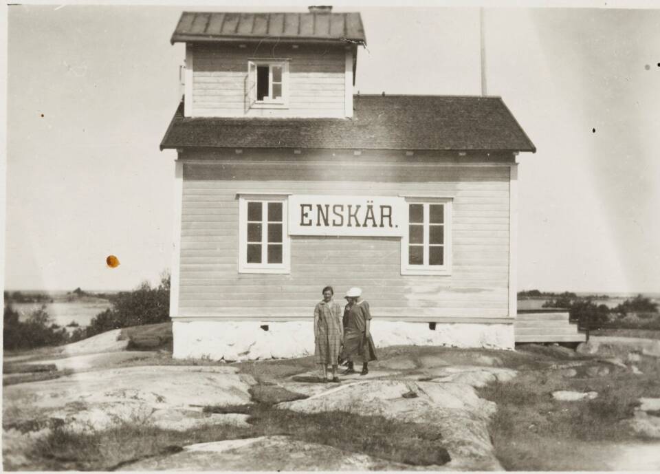 Vanhassa huonokuntoisessa mustavalkokuvassa puutalon (seinässä lukee Enskär) edustalla kolme naishenkilöä pitkissä mekoissa, talo kallion laella.