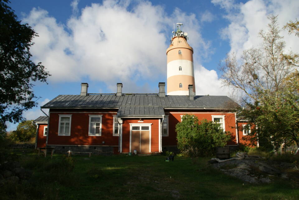 Aurinko paistaa matalaan pitkähköön puutaloon, jonka takaa nousee kaksivärinen majakka.