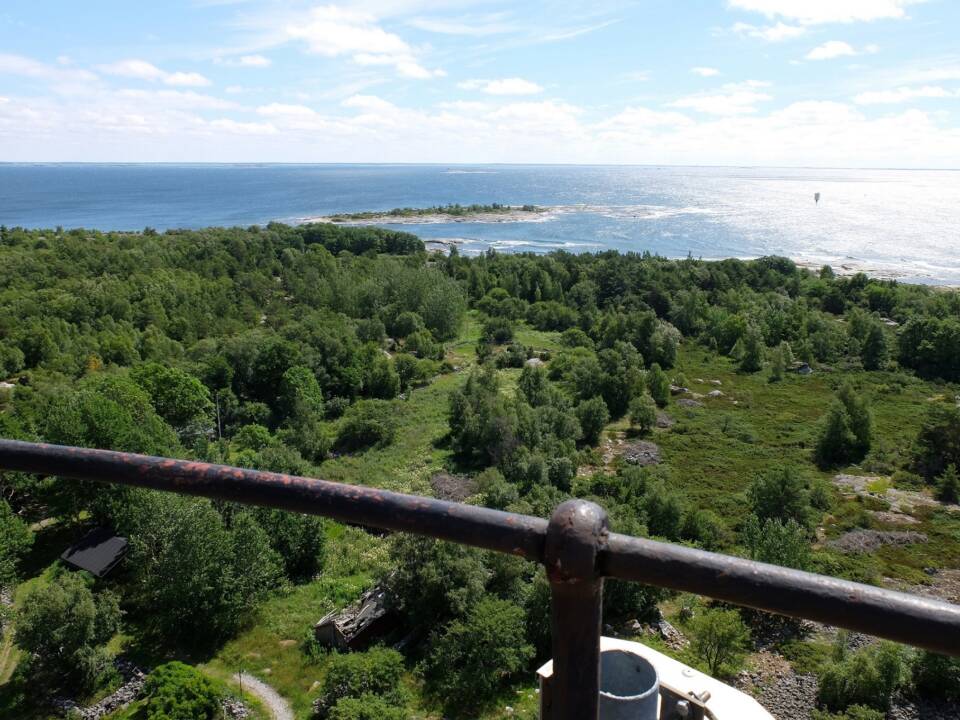 Majakan kaiteen yli katsotaan metsäistä saarta, jonka takaa alkaa aurinkoinen meri.
