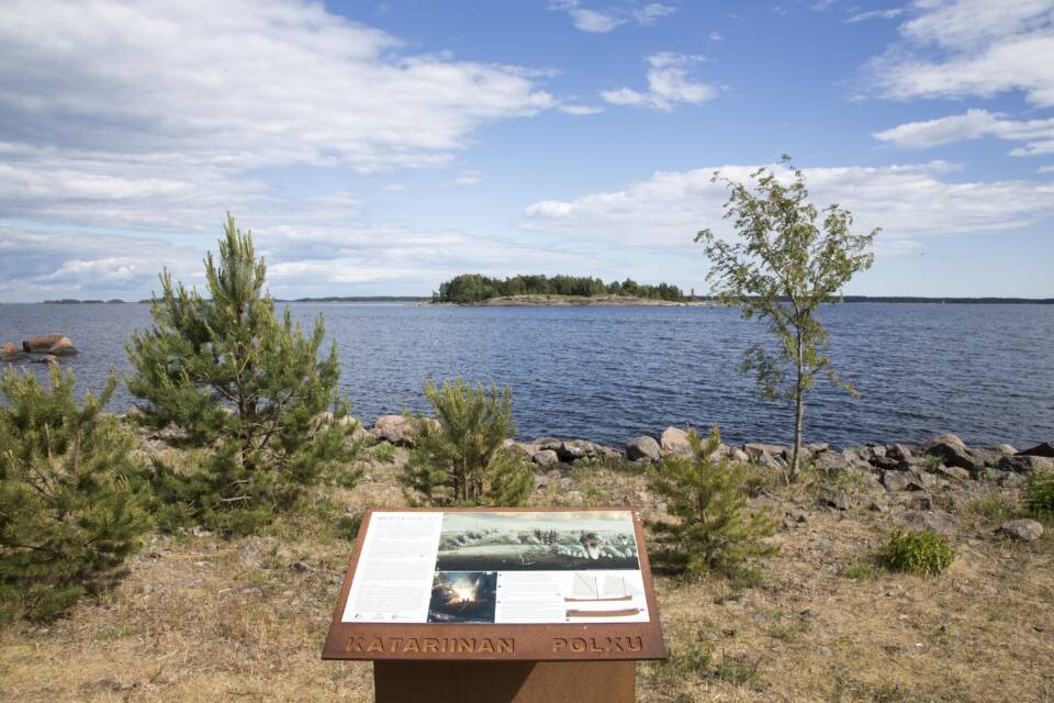 Hiekkaiselta maalta katsotaan merelle ja siellä olevaan saareen, etualalla infokyltti merelle päin.