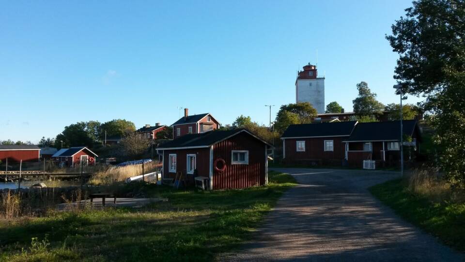 Punamultaisia puisia rakennuksia majakan edustalla, kylänraitti, aurinkoinen kesäpäivä.