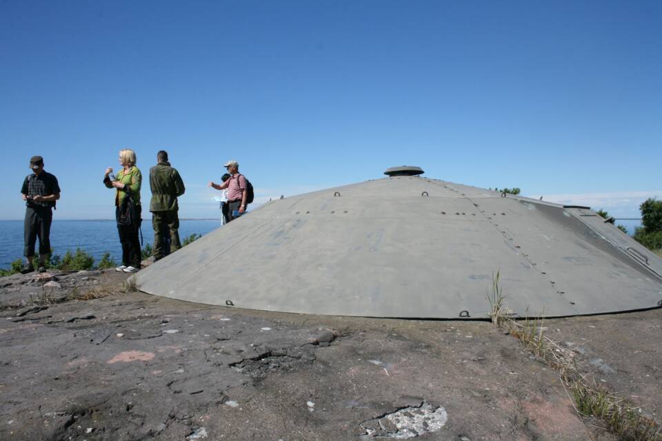 Kallion laella seisoo ihmisiä katselemassa merellä, heidän takanaan lähes ihmisten korkuinen metallinen/betoninen pyöreä koroke.