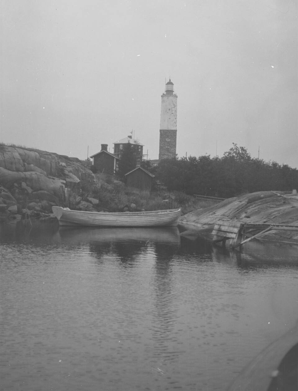 Vanhassa mustavalkoisessa valokuvassa etualalla kalliolahti ja vene, kalliolla metsän reunassa rakennuksia, taustalla majakka.