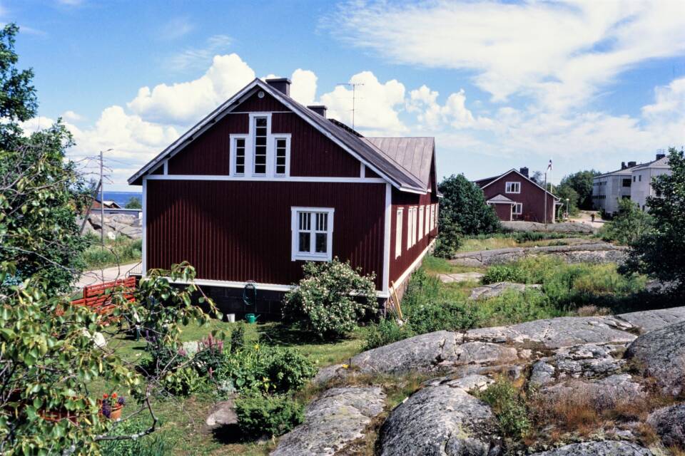 Pitkä ja lyhyempi punainen puutalo aurinkoisella ja lehtipuisella saarella.