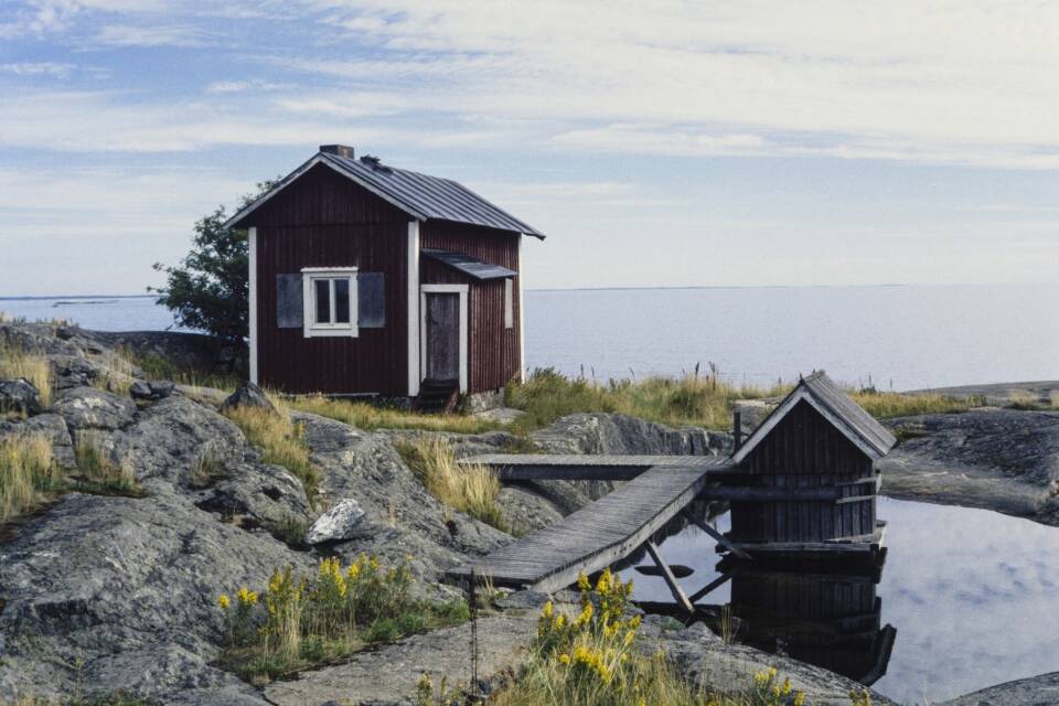 Punamultainen puutalo, laituri ja katettu kaivonkansi kalliosaarella.