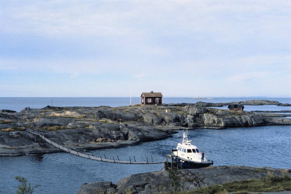 Kaksi kalliosaarta, joita yhdistää riippusilta, lähemmän saaren rannassa vene, kauemmalla saarella talo.