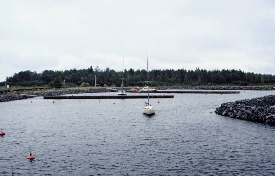Kaksi purjevenettä ja moottorivenettä muuten autiossa suojasatamassa, taustalla puustoinen saari ja rakennuksia.