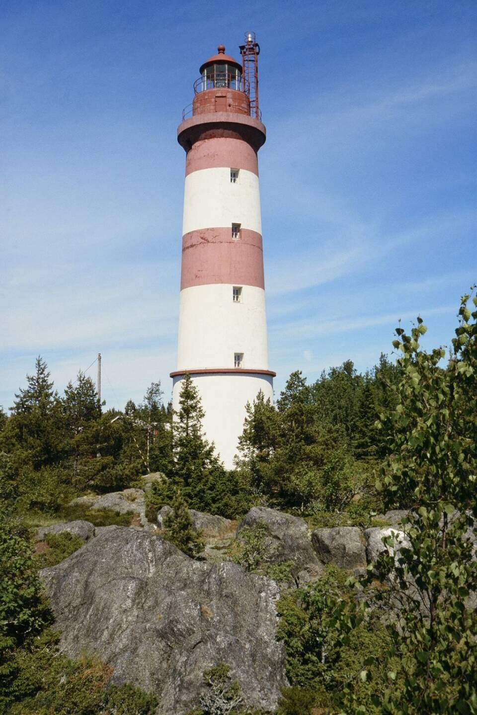 Kaksivärinen majakka seisoo kalliolla matalien lehtipuiden ympäröimänä, sininen pilvetön taivas.