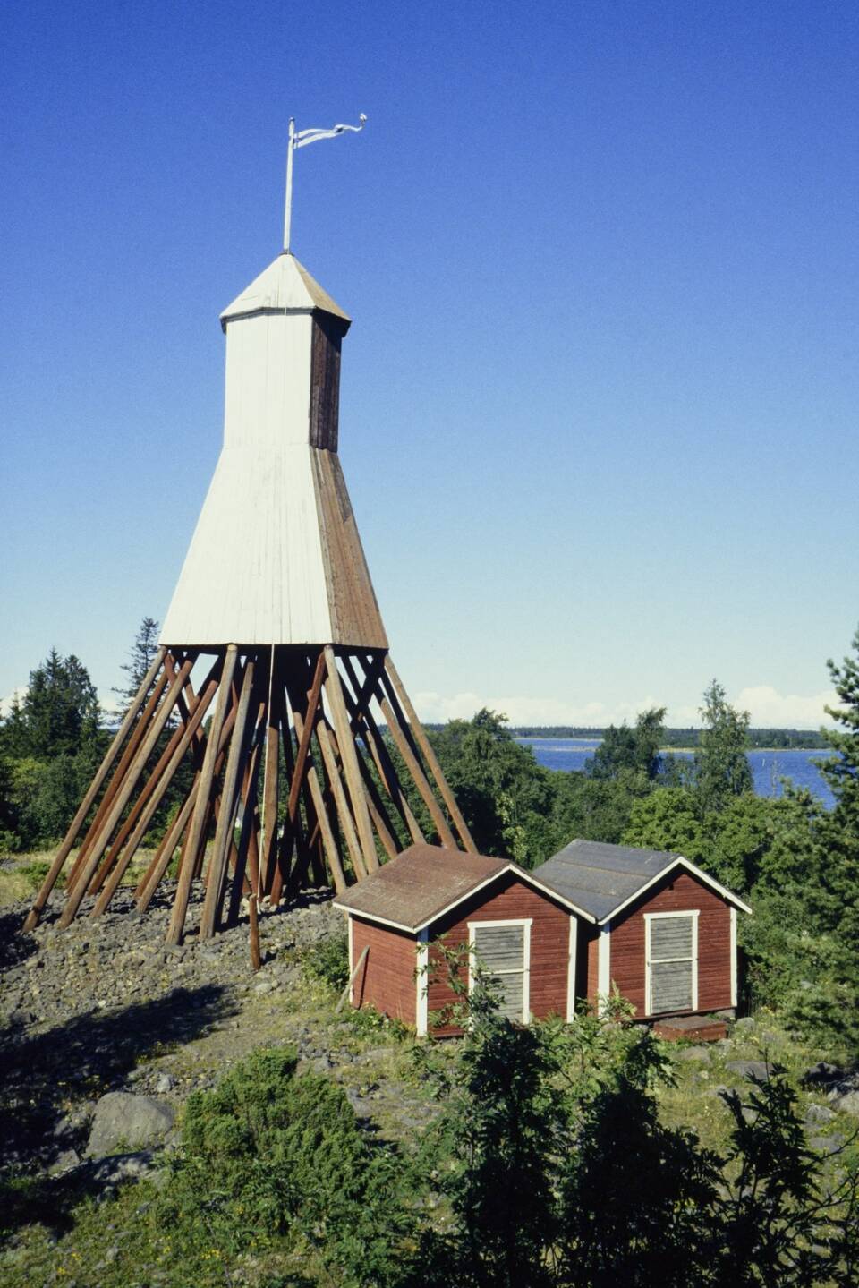 Kartionmallinen, kuin puujaloilla seisova majakka, sen edustalla puustoisella kalliosaarella kaksi pientä aittaa.