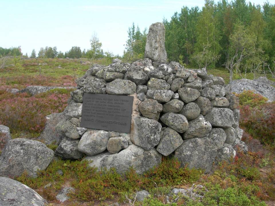 Suurista pyöreähköistä kivistä rakennettu keko, jossa muistolaatta, varvikkoisella ja kivikkoisella nummella.