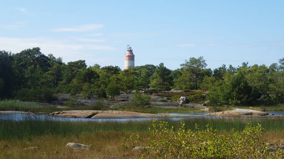 Majakka metsän takana saarella lahden yli katsottuna, etualalla niitty.