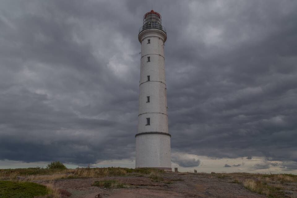 Yksinkertainen, yksivärinen, pyöreä majakka kalliosaaren laella, taustalla dramaattiset tummat pilvet.