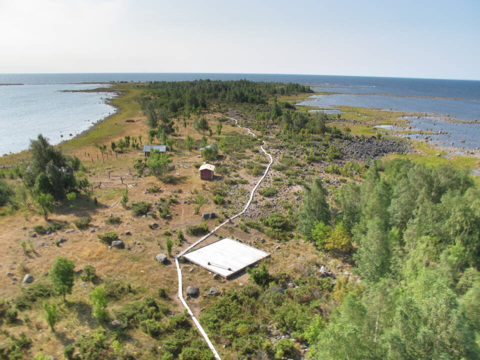 Kapea matala ja osittain puustoinen saari, jonka toiseen päähän vie pitkospuut saaren keskeltä, helikopterin laskeutumislava, rannat matalat ja rikkonaiset, tornista nähtynä.